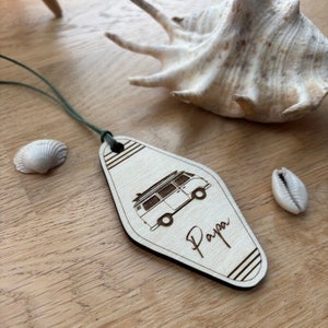 Peut inclure: Porte-clés en bois avec un design de van vintage et le mot "Papa" gravé dessus. La étiquette est attachée à un cordon vert.