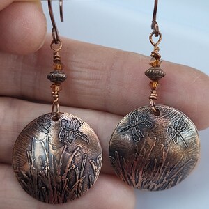 May include: A pair of copper earrings with a dragonfly and grass design. The earrings are accented with small amber-colored beads.