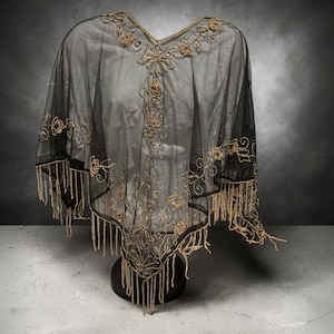 Peut inclure: Un poncho en maille noire transparente orné de perles dorées complexes et de broderies à motifs floraux le long de l'encolure et du corps. Le vêtement est fini avec une bordure à franges dorées décoratives le long de l'ourlet inférieur.