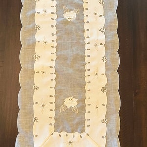 May include: White table runner with a scalloped edge and embroidered floral designs. The runner has a delicate, lacy look and would add a touch of elegance to any table setting.