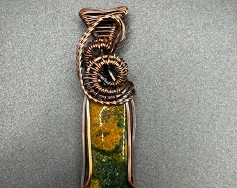 May include: A handcrafted pendant featuring a rectangular stone with green and gold hues, encased in copper wire. The wirework includes intricate coils and a bail for a necklace. The pendant is approximately 2 inches long.