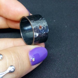 May include: A wide, hammered silver ring with a single, small, round, orange-brown gemstone. The ring has a textured surface and a dark, oxidized finish. The ring is held up against a black background.