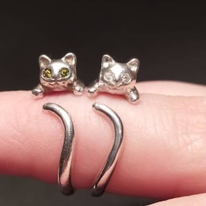 Può includere: Due anelli d'argento con disegni di gatti. I gatti sono rivolti in avanti con occhi verdi e le loro code sono avvolte attorno al dito.