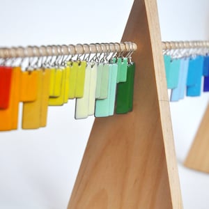 Puede incluir: Una colección de pendientes rectangulares en un espectro completo de colores, incluyendo rojo, naranja, amarillo, verde, azul y morado, colgados de una barra de madera horizontal sostenida por dos soportes de madera triangulares.