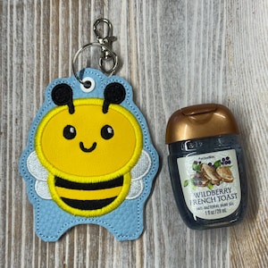 May include: A light blue bee-shaped embroidered keychain holder with a metal clasp and a small bottle of Wildberry French Toast anti-bacterial hand gel. The hand gel bottle is clear with a bronze cap and a label stating 1 fl oz/29 ml.