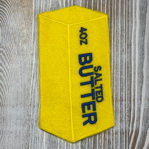 May include: A bright yellow felt patch shaped like a rectangular stick of butter. The patch features embroidered blue text that reads 4OZ SALTED BUTTER. The design includes stitched lines to create a three-dimensional appearance of a butter stick.