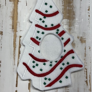 Peut inclure: Ornement de sapin de Noël en feutre blanc, avec des détails rouges et verts. La forme de l'arbre présente une ouverture centrale et est ornée de lignes courbes rouges et de petits carrés verts. Conçu pour être suspendu.