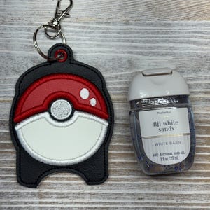 May include: A black faux leather hand sanitizer holder featuring an embroidered red and white ball design with a silver metal lobster clasp keychain, positioned next to a clear plastic bottle of Fiji White Sands anti-bacterial hand gel.