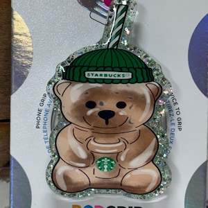 May include: A brown bear-shaped PopGrip phone accessory, holding a Starbucks coffee cup. The bear wears a green beanie with the Starbucks logo. The grip has a glittery border and the text "POPGRIP" is visible.