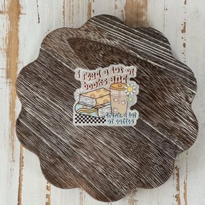 May include: A brown, wooden, scalloped-edge coaster with a sticker that reads "I read a lot of books and drink a lot of coffee." The sticker features illustrations of books, a coffee drink, and a daisy.