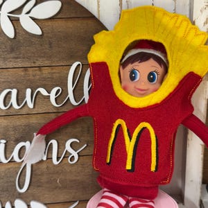 May include: An elf doll wearing a red and yellow felt costume resembling a fast-food french fry container. The costume has a yellow collar and a red top with a yellow "M" logo. The elf has a red and white striped legs.