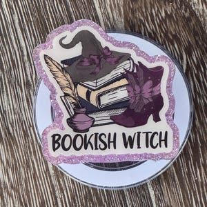 May include: A round, white object with a glittery purple border featuring a "Bookish Witch" design. The design includes a witch hat, books, a feather quill, an inkwell, and a purple boot. The overall theme is fantasy and reading.