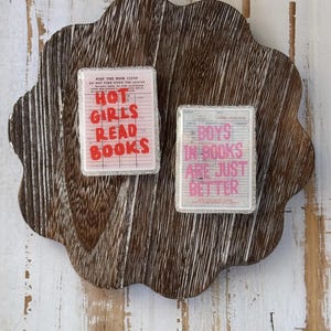 May include: Two rectangular book-themed decorative items with text on a wooden tray. One item reads "HOT GIRLS READ BOOKS" in red letters. The other reads "BOYS IN BOOKS ARE JUST BETTER" in pink letters. Both items have a silver glitter border.