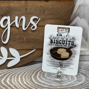 Può includere: Un porta badge con la scritta "Some of y'alls BISCUITS ain't DONE IN THE MIDDLE" e una grafica di biscotti in una padella. Il porta badge è su una carta bianca con istruzioni per la cura. Lo sfondo è un cartello di legno.