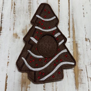 May include: A brown felt Christmas tree ornament with white and red embroidered details. The tree has a circular opening in the center and is decorated with white stripes and red dots. The ornament is designed to look like a gingerbread cookie.