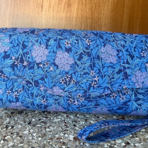 May include: A rectangular fabric clutch bag featuring a vibrant blue and light purple floral pattern with dense foliage and small blossoms. The accessory includes a fold-over flap closure and a matching fabric wrist strap attached to the side.
