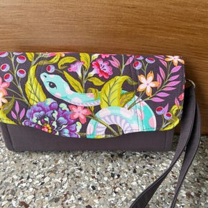 May include: A rectangular clutch bag with a dark gray base and a flap featuring a colorful print of a light blue snake winding through vibrant pink, purple, and orange flowers with green leaves. The bag includes a matching dark gray wrist strap.