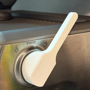 Puede incluir: Primer plano de una palanca blanca en una máquina de espresso de acero inoxidable. La palanca está situada encima del botón "STANDBY" y presenta un diseño curvo. La máquina tiene iconos de vapor y de gota de agua.
