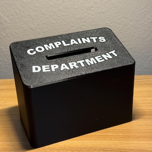 May include: A black rectangular box with a slot on top, labeled "COMPLAINTS DEPARTMENT" in white capital letters. The box is sitting on a wooden surface.