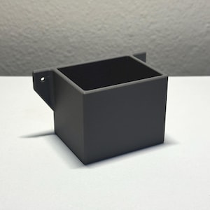 May include: A small, gray, rectangular container with a square opening. The container has a small, angled bracket with a hole on one side. The interior of the container is black. The object is sitting on a white surface.