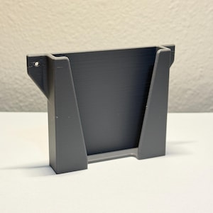 Puede incluir: Un soporte de teléfono de pared gris impreso en 3D. El soporte tiene una parte trasera rectangular con dos soportes angulados y un pequeño labio en la parte inferior. Un pequeño agujero es visible en el lado izquierdo. El soporte está diseñado para sostener un teléfono u otro dispositivo pequeño.