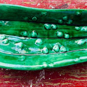 May include: A glossy, elongated ceramic dish shaped like a pickle, featuring a vibrant emerald green glaze. The surface is textured with raised, rounded bumps that mimic the natural appearance of a cucumber, with a deep central groove.