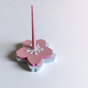 May include: A pink and white flower-shaped desk organizer with a central pink rod. The organizer has a layered design with a light blue base, a pink middle layer, and a pink top layer with white accents.