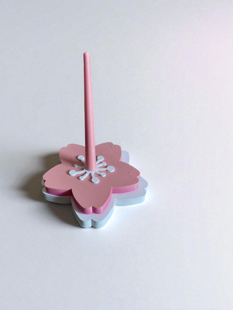 May include: A pink and white flower-shaped desk organizer with a central pink rod. The organizer has a layered design with a light blue base, a pink middle layer, and a pink top layer with white accents.