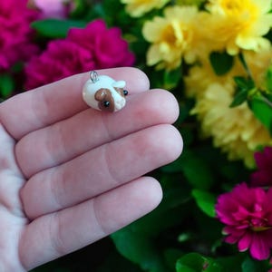 Puede incluir: Un pequeño dije artesanal de cobaya. El dije es blanco con marcas marrones y ojos negros. Tiene un lazo plateado en la parte superior para sujetarlo a un collar o llavero. El fondo presenta flores coloridas.