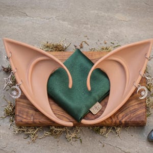 Puede incluir: Un par de orejas de elfo color carne con colgantes plateados de estrella y luna, sobre una tabla de madera con un cuadrado de tela verde. Cerca hay una amatista púrpura y una piedra azul gris.
