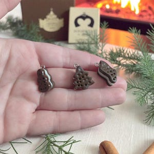 Puede incluir: Tres dijes en miniatura de color chocolate con forma de galleta, árbol de Navidad y galleta. Cada dije tiene un lazo plateado en la parte superior. Los dijes se sostienen en una mano, con un fondo borroso de una chimenea y decoraciones navideñas.