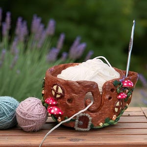 May include: A whimsical brown yarn bowl with a fairy house design, featuring red and white mushroom accents. The bowl holds white yarn, with a crochet hook inserted. Two balls of yarn, one teal and one pink, sit beside the bowl on a wooden surface.