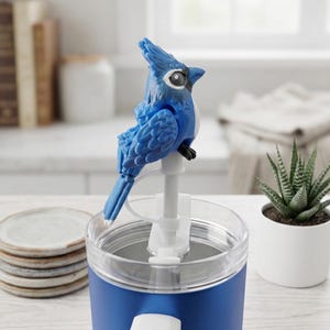 May include: A blue and white bird-shaped bottle stopper is mounted on a blue and clear container. The bird has detailed feather textures and a white eye. A small potted succulent plant and a stack of plates are in the background.