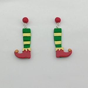 May include: A pair of green and yellow striped elf leg earrings with red shoes and a gold bell on the toe. The earrings are made of polymer clay and have a silver hook.