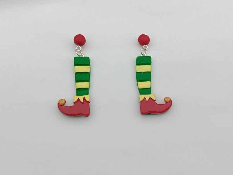 May include: A pair of green and yellow striped elf leg earrings with red shoes and a gold bell on the toe. The earrings are made of polymer clay and have a silver hook.
