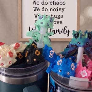 Op de afbeelding: Een verzameling kleurrijke, grillige draakvormige dranktoppers in verschillende kleuren, waaronder groen, blauw, roze en crème. Elke draak zit bovenop een doorzichtige rietje. Op de achtergrond staat: "We do chaos, We do noisy, We do hugs, love, family".