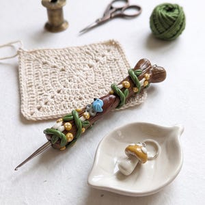 May include: A brown polymer clay crochet hook handle decorated with green vines, small yellow mushrooms, and a tiny blue bird. Beside it sits a white leaf-shaped ceramic dish containing a mushroom-shaped stitch marker with a silver metal ring.