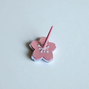 May include: A pink flower-shaped object with a matching pink stem. The flower has a white outline and white details in the center. The object is likely a decorative item or a small accessory.