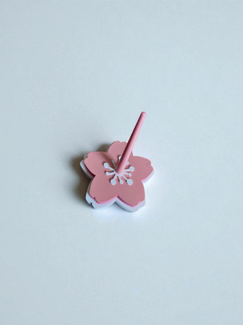 May include: A pink flower-shaped object with a matching pink stem. The flower has a white outline and white details in the center. The object is likely a decorative item or a small accessory.