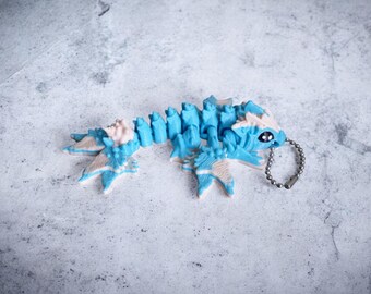 Pode incluir: Um chaveiro de criatura articulada azul e branca com uma corrente prateada. O brinquedo tem um corpo segmentado e detalhes, incluindo um pequeno olho. O esquema de cores é uma mistura de azul claro, branco e um toque de rosa.