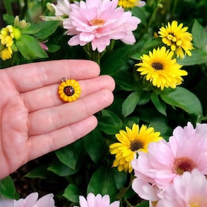 May include: A small yellow sunflower charm with a brown center, held in a hand against a background of pink and yellow flowers.