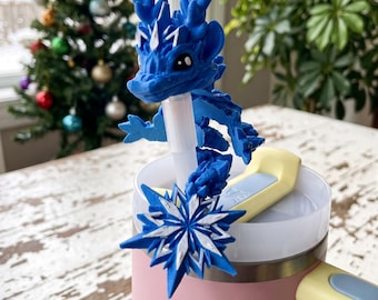 May include: A blue 3D-printed dragon straw topper featuring a white snowflake accent. The dragon has detailed scales, horns, and large dark eyes. It is attached to a clear straw inserted into a pink tumbler with a yellow and light blue handle.