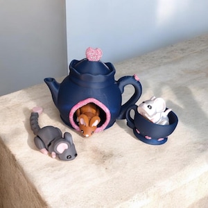 Puede incluir: Una tetera azul con un corazón rosa en la parte superior, un ratón marrón dentro y un ratón blanco en una taza de té azul con detalles rosas.