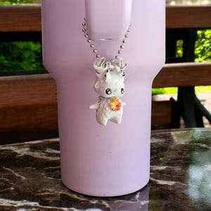 May include: A white deer-like pendant with a silver chain hangs in front of a light purple tumbler. The pendant has small antlers, black eyes, and a colorful flower-like design on its chest. The tumbler is on a marble-like surface.