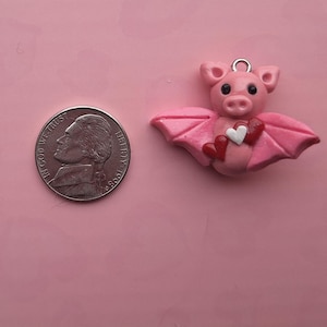 May include: A pink bat-pig charm with a silver loop for hanging, holding a white and red heart. A nickel coin is next to the charm on a pink background. The charm has black eyes and pink wings.