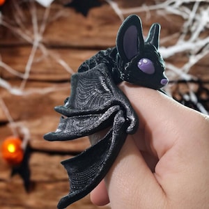 Puede incluir: Una figurita de murciélago negro impresa en 3D con ojos morados. El murciélago está posado con las alas plegadas y está siendo sostenido por una mano.