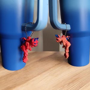 Könnte beinhalten: Zwei blau-verlaufende Becher mit roten Drachenanhängern, die an den Griffen hängen. Die Drachenanhänger haben blaue Akzente. Die Becher stehen auf einer hellbraunen Holzoberfläche.