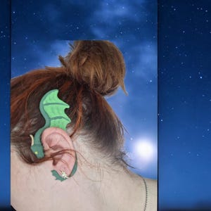 以下が含まれることがあります： 緑色のドラゴン型のイヤーカフで、黄色の目と金の星のチャームが付いています。イヤーカフは人の耳につけられています。