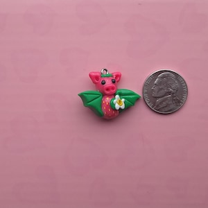 May include: A handmade pink and green dragon charm with a white flower and a silver-colored loop. The dragon has green wings and a strawberry-like body. A US nickel is next to the charm for size comparison.
