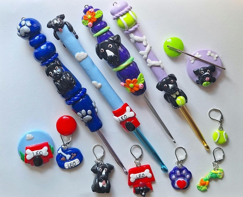 Op de afbeelding: Een set van 10 haaknaalden met hondenthema toppers. De toppers zijn gemaakt van polymer clay en hebben zwarte honden, botten, tennisballen en voetafdrukken. De haaknaalden zijn in verschillende kleuren, waaronder blauw, paars en goud. De haaknaalden zijn gelabeld "Leo" en hebben een hondenhok ontwerp.